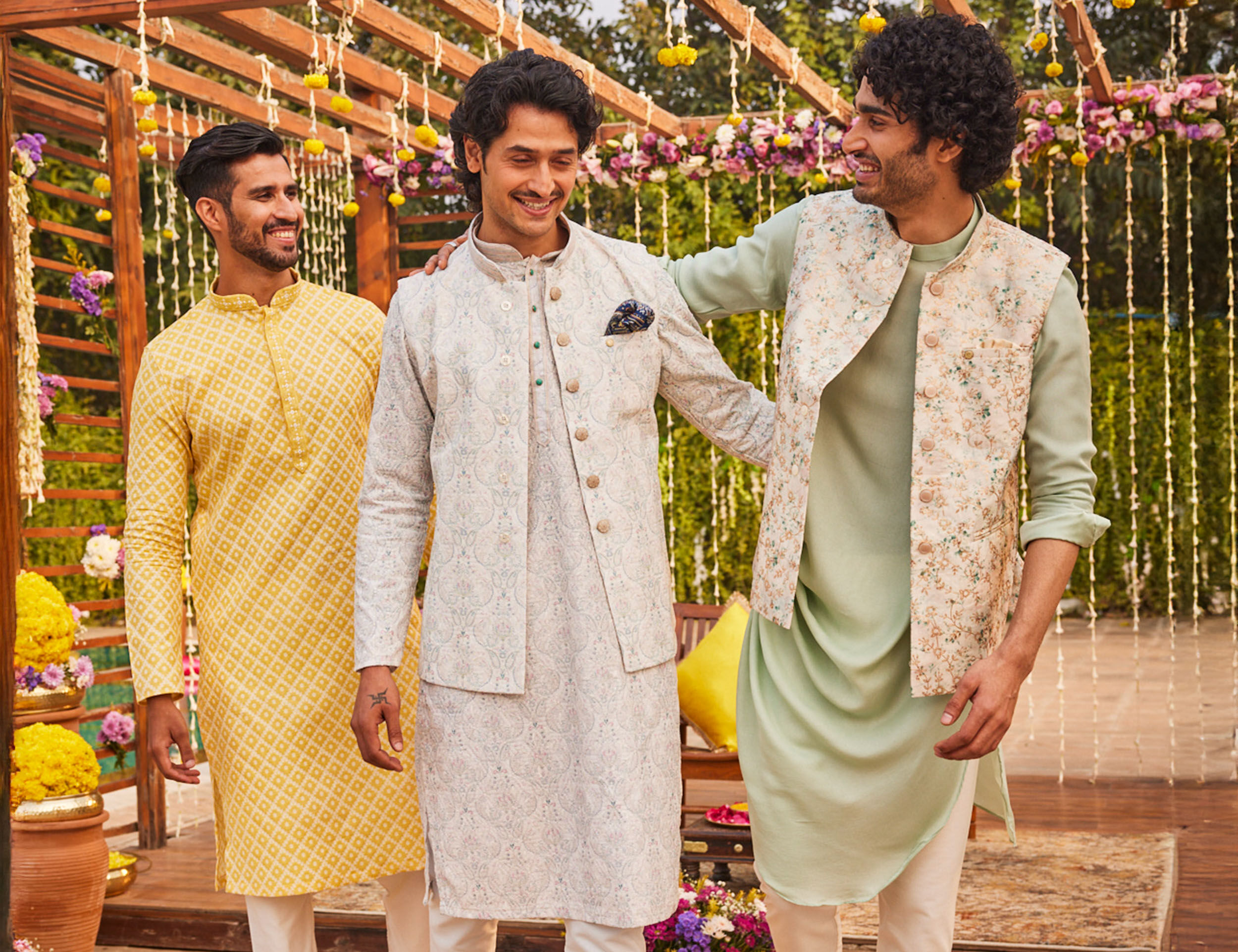 Three men wearing stylish kurta pajama sets in pastel and festive tones, posing together at a floral-decorated outdoor celebration setup